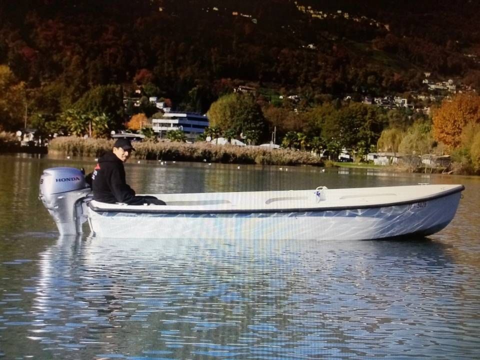 Persona a bordo di una piccola barca a motore su un lago tranquillo, con alberi autunnali sullo sfondo.
