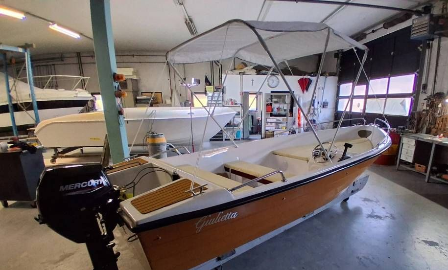 Piccola barca a motore in legno su un rimorchio all'interno di una rimessa per barche, con tendalino bianco e scafo color sabbia.