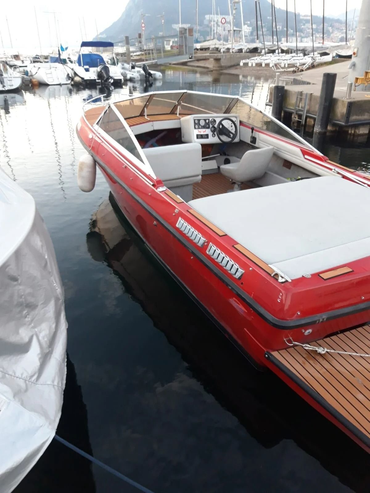 Motoscafo rosso ormeggiato in un porticciolo accanto ad acque calme e barche a vela, con le montagne sullo sfondo.