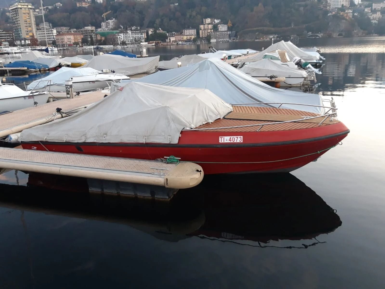 Ti Yachts Sagl
Barca rossa coperta da teloni bianchi ormeggiata su acque calme vicino a una costa innevata e a case lontane.