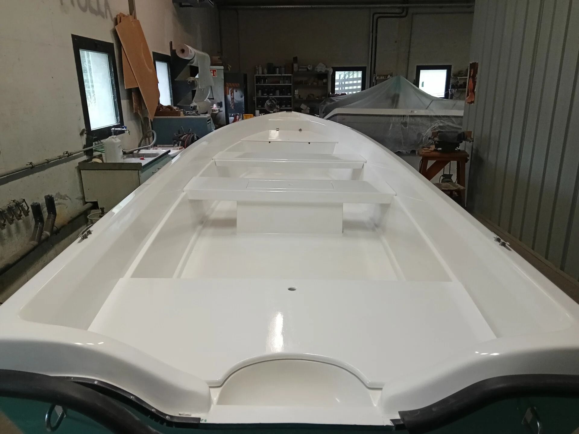 Scafo bianco di una barca in un'officina, visto da poppa all'interno di uno spazio industriale simile a un garage.