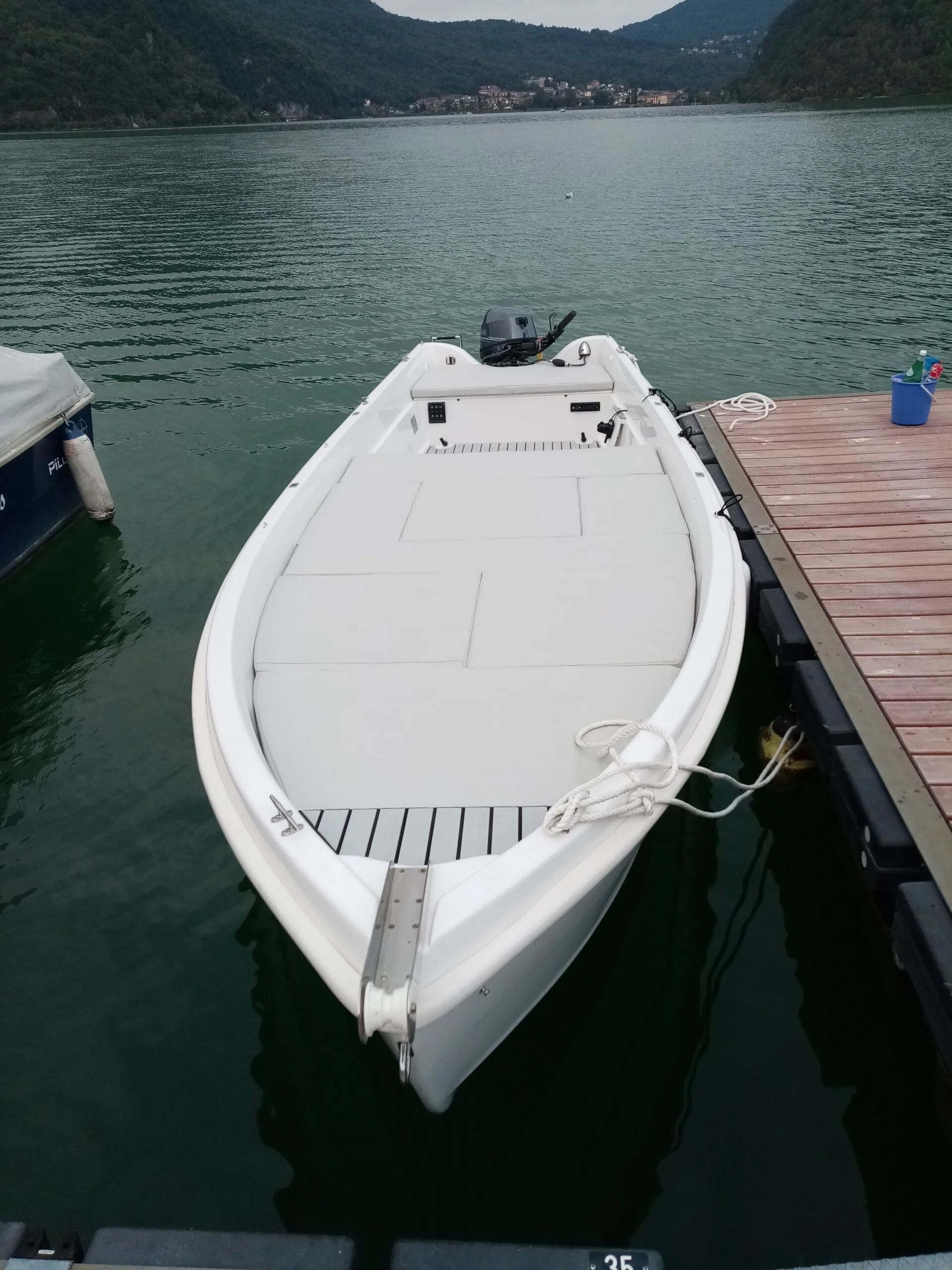 Motoscafo bianco ormeggiato accanto a un molo di legno su acque verdi e calme, con montagne sullo sfondo.