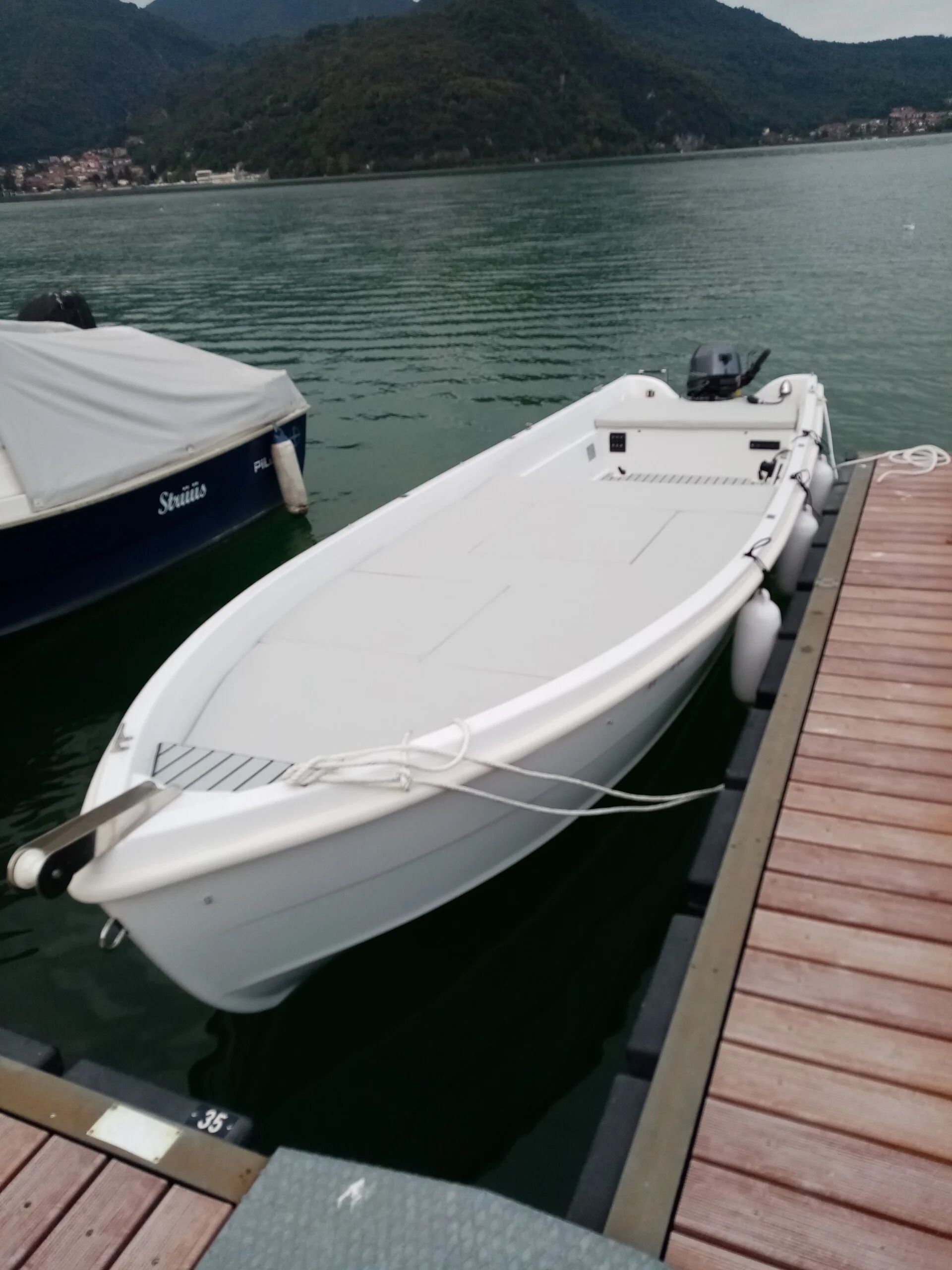 Motoscafo bianco ormeggiato a un molo su acque calme, con montagne sullo sfondo.