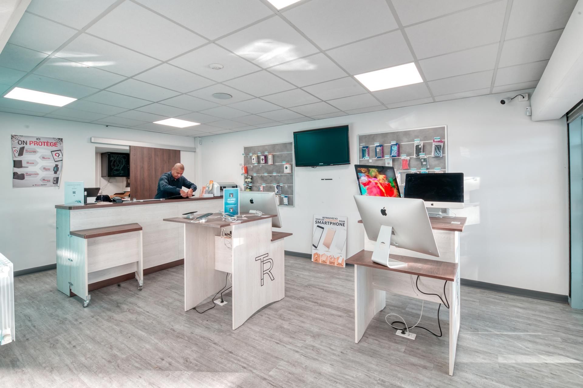 Un atelier de réparation électronique propre et bien éclairé, avec un comptoir de service, deux postes de travail informatiques et des présentoirs muraux.