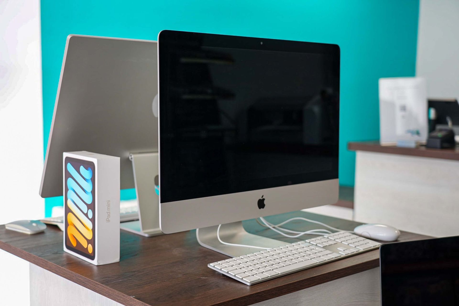 Un bureau avec un iMac d'Apple, un clavier blanc, une souris et une boîte d'iPad mini, le tout adossé à un mur turquoise.
