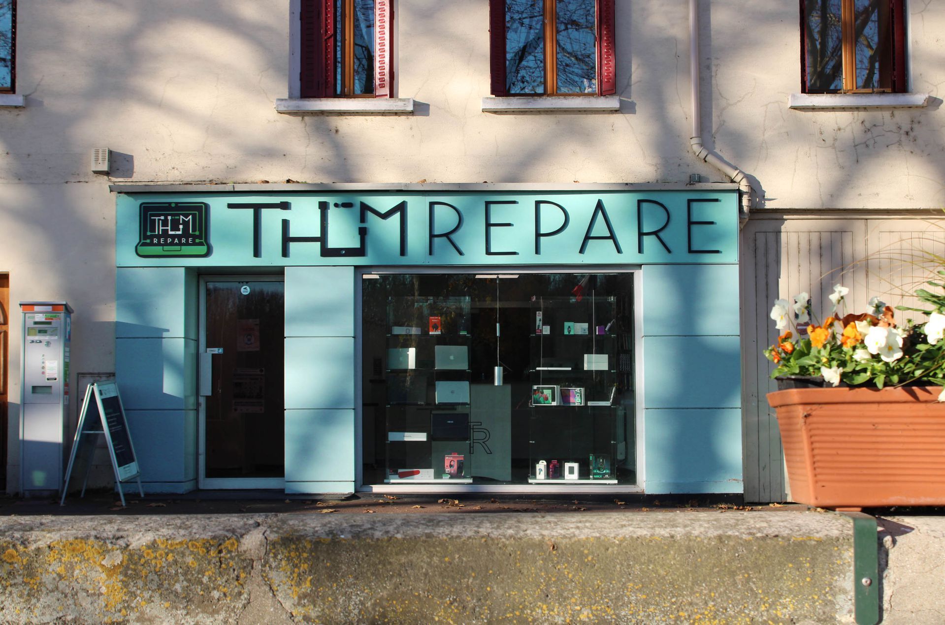 Une devanture aux murs bleu clair et l'enseigne THUM REPARE au-dessus d'une vitrine exposant des appareils électroniques.