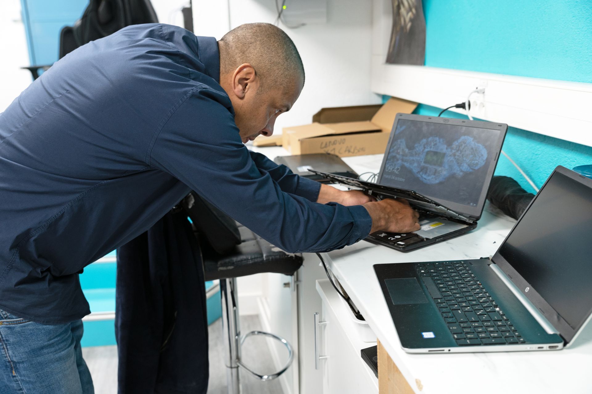 Une personne en chemise bleue penchée sur un bureau pour réparer un ordinateur portable ouvert, un autre ordinateur portable se trouvant à proximité sur le bureau.