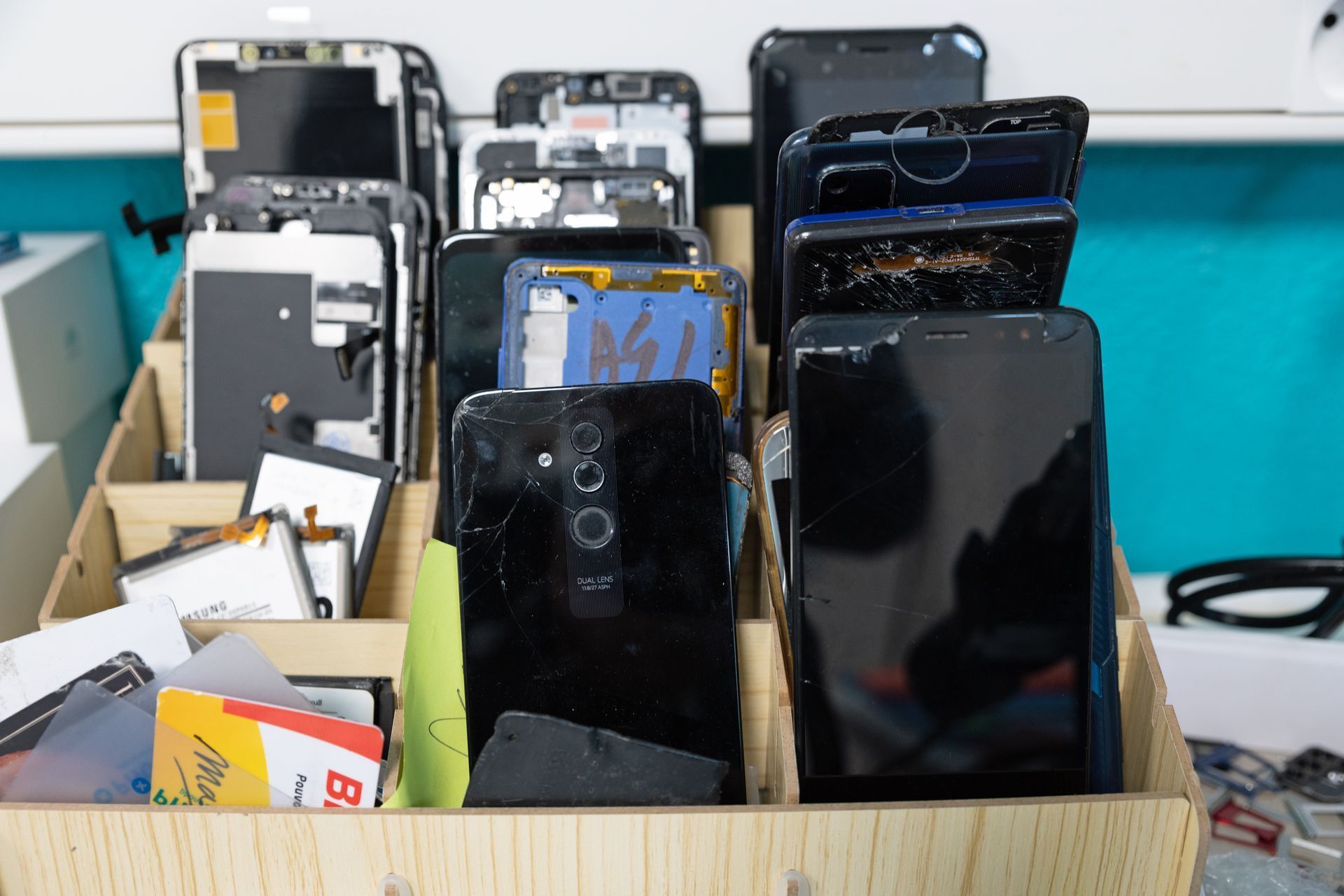 Une caisse de rangement en bois remplie de divers smartphones démontés et de composants électroniques, sur un fond bleu clair.