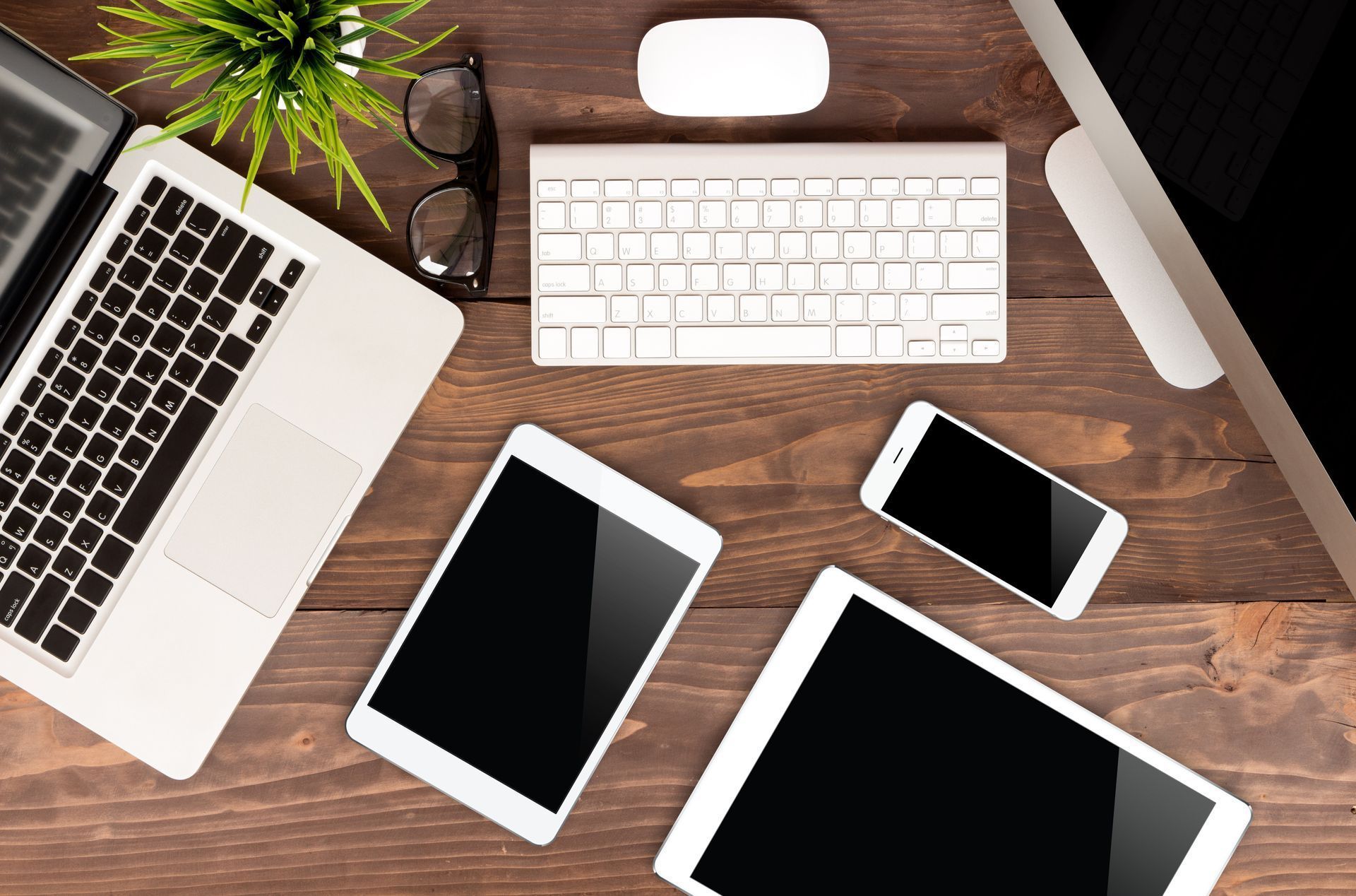 Vue en plongée d'un bureau montrant un ordinateur portable, un clavier, une souris, deux tablettes, un téléphone et une petite plante en pot.