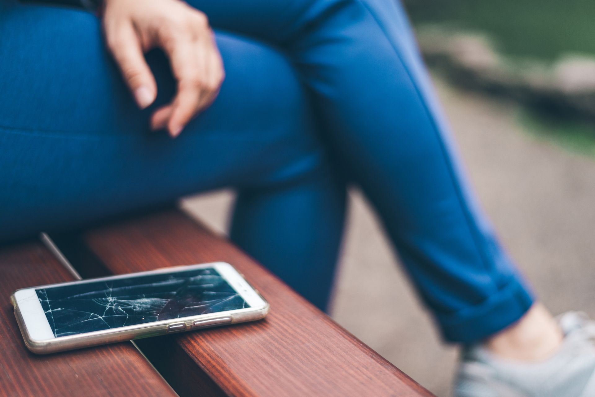 Un smartphone à l'écran brisé est posé sur un banc en bois, à côté d'une personne portant un pantalon bleu.
