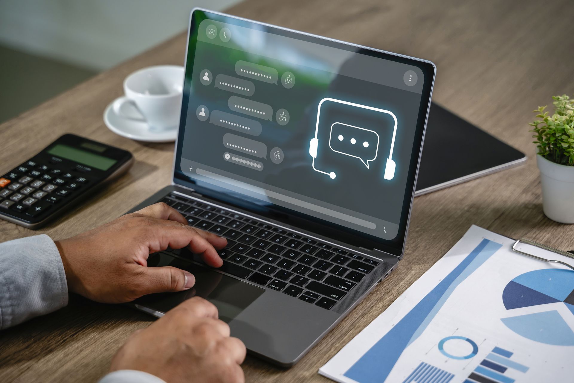 Une personne tape sur un ordinateur portable affichant une interface de chat d'assistance client, posée sur un bureau en bois avec une calculatrice et des graphiques.