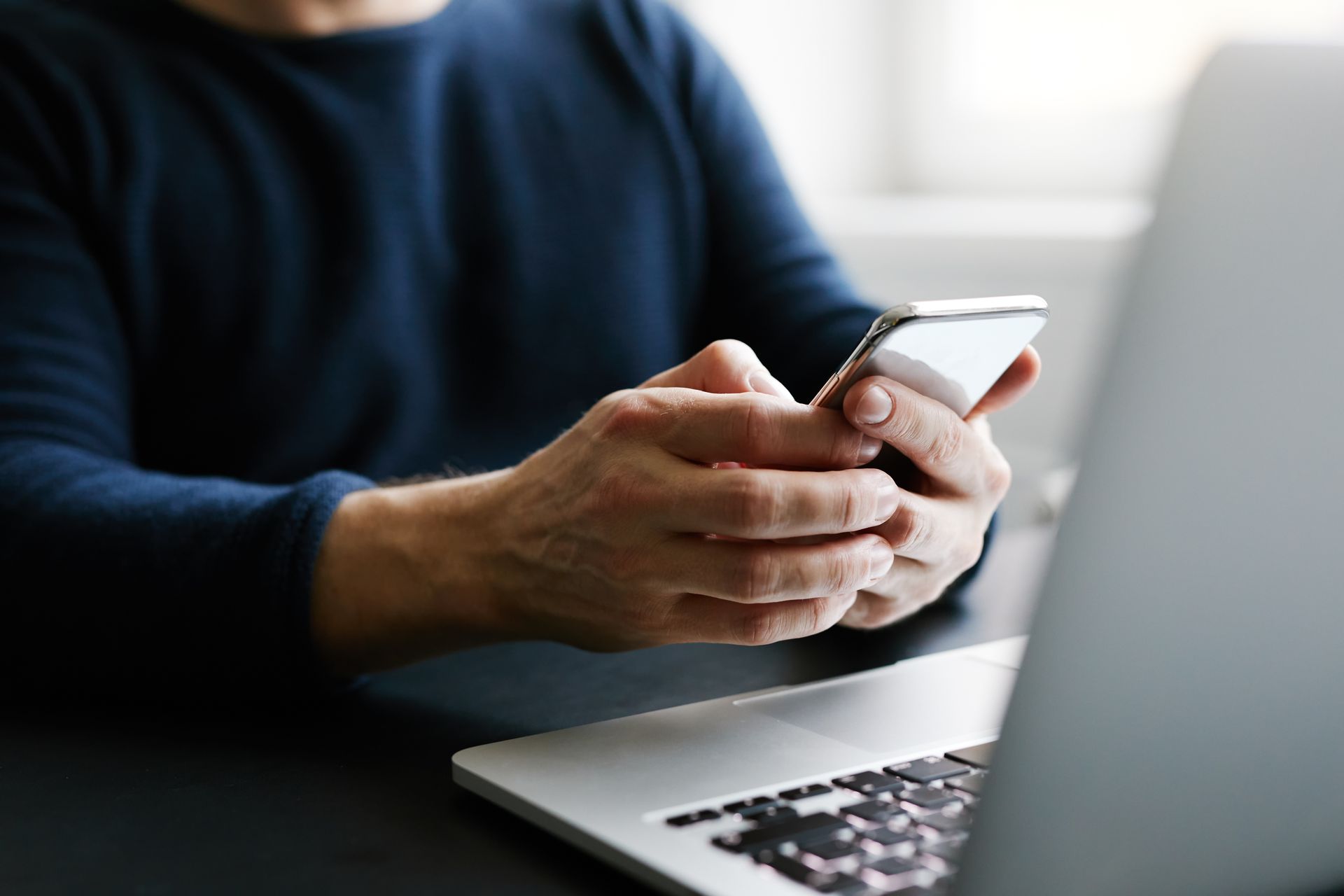 Une personne portant un pull bleu utilise un smartphone tout en étant assise devant un ordinateur portable.