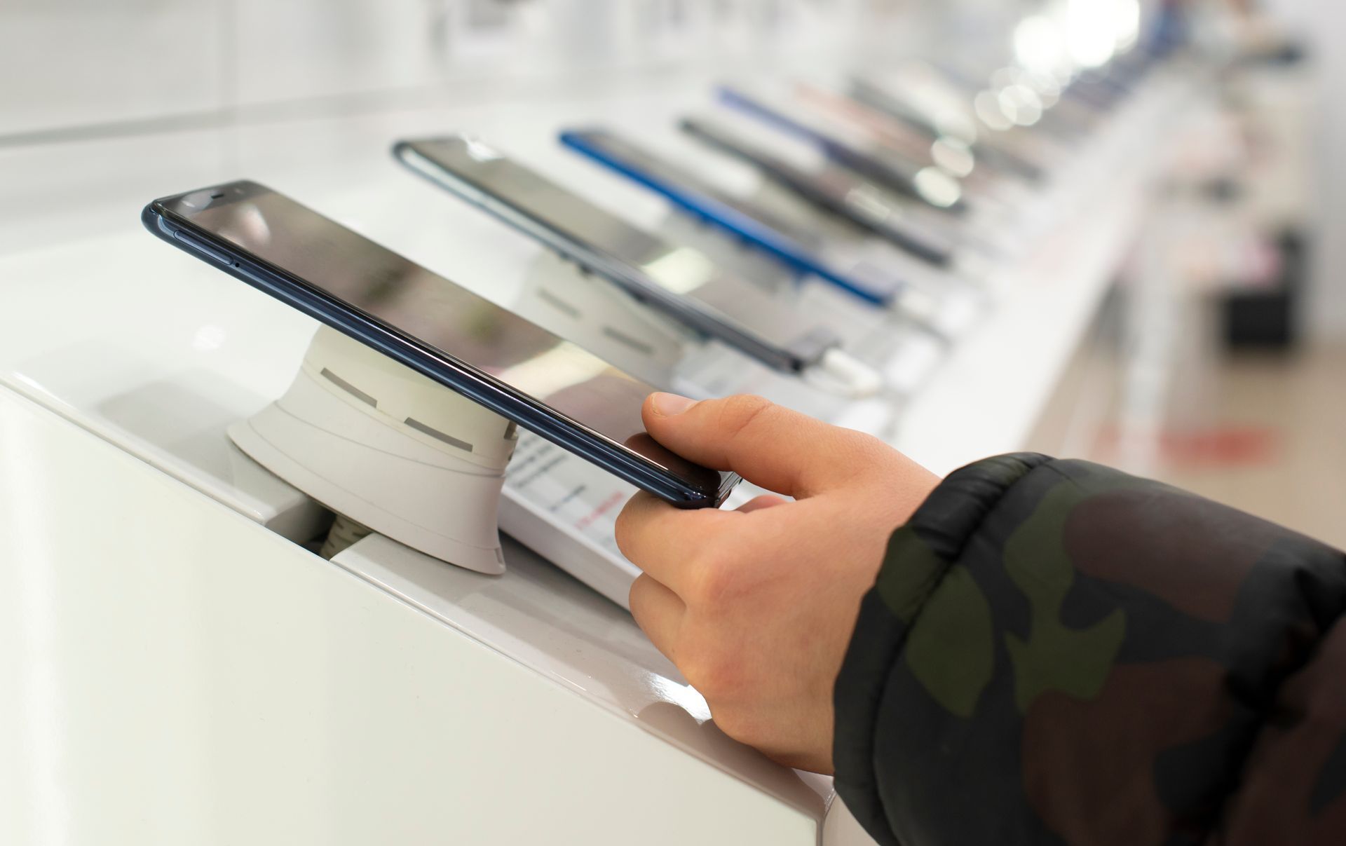 Une main effleure un smartphone exposé sur un présentoir dans un magasin d'électronique très éclairé, avec d'autres téléphones en arrière-plan.