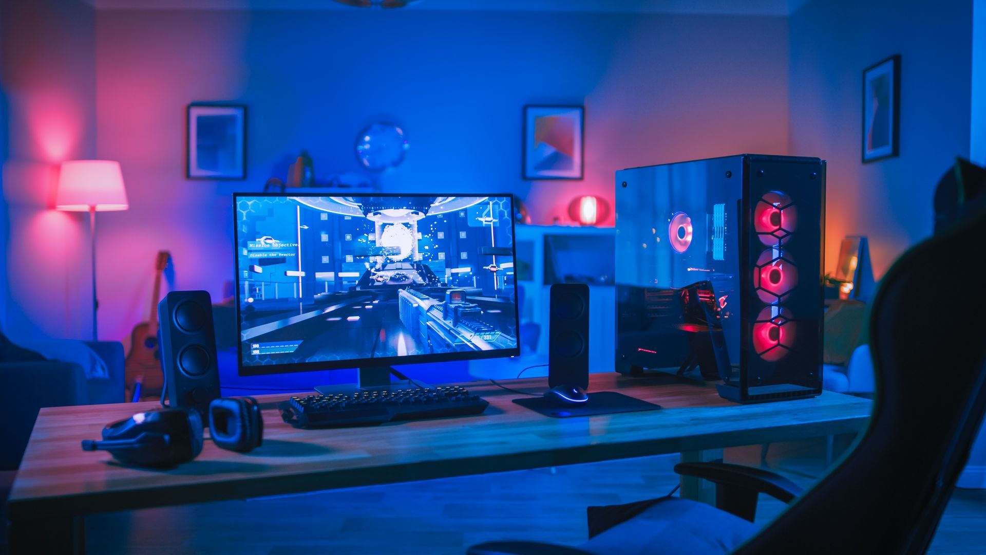 Une pièce faiblement éclairée avec un PC de jeu, un écran et des périphériques sur un bureau en bois, baignée d'une lumière ambiante bleue et rouge.