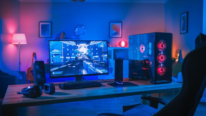 Un poste de jeu avec un écran affichant un jeu, une tour d'ordinateur avec des lumières rouges et des périphériques sur un bureau dans une pièce bleue.
