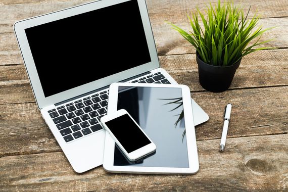 Un ordinateur portable, une tablette et un smartphone sont disposés sur une table en bois rustique, à côté d'une petite plante en pot et d'un stylo blanc.