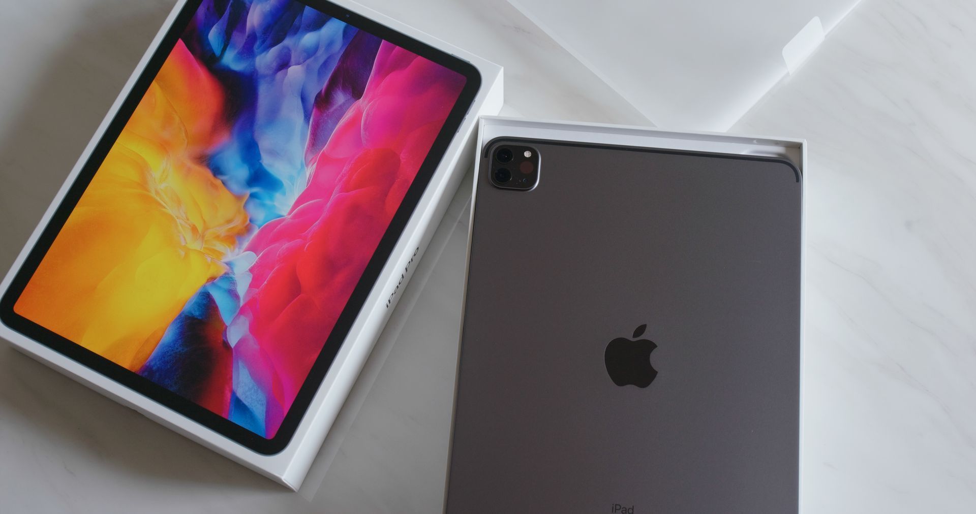 Un iPad Pro gris posé à côté de sa boîte, qui affiche un fond d'écran abstrait aux couleurs vives.