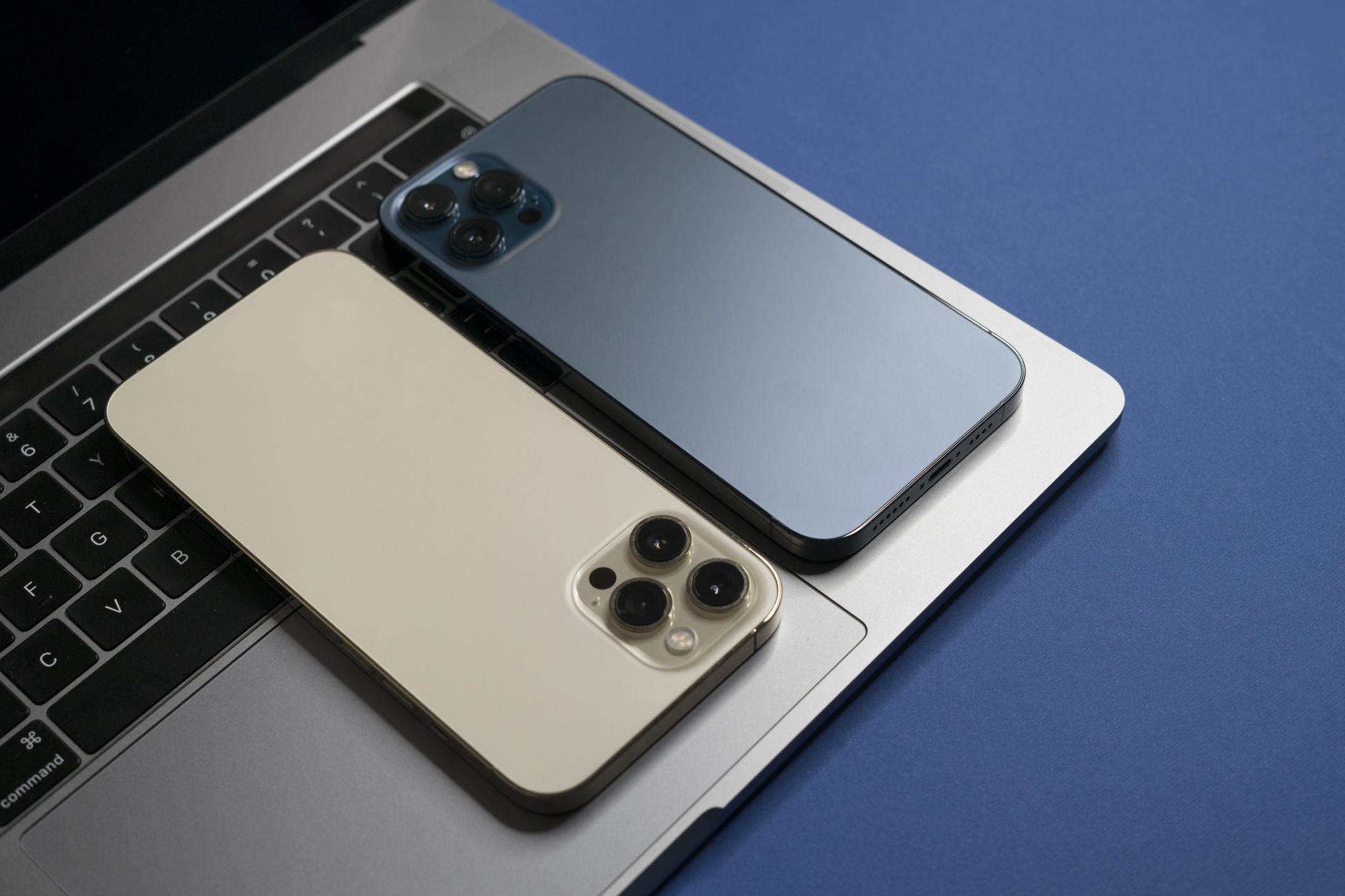 Deux smartphones, l'un doré et l'autre bleu foncé, posés sur un ordinateur portable argenté ouvert, sur fond bleu.