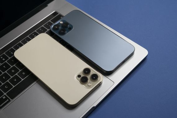 Deux smartphones, l'un doré et l'autre bleu foncé, posés sur un ordinateur portable argenté ouvert, sur fond bleu.