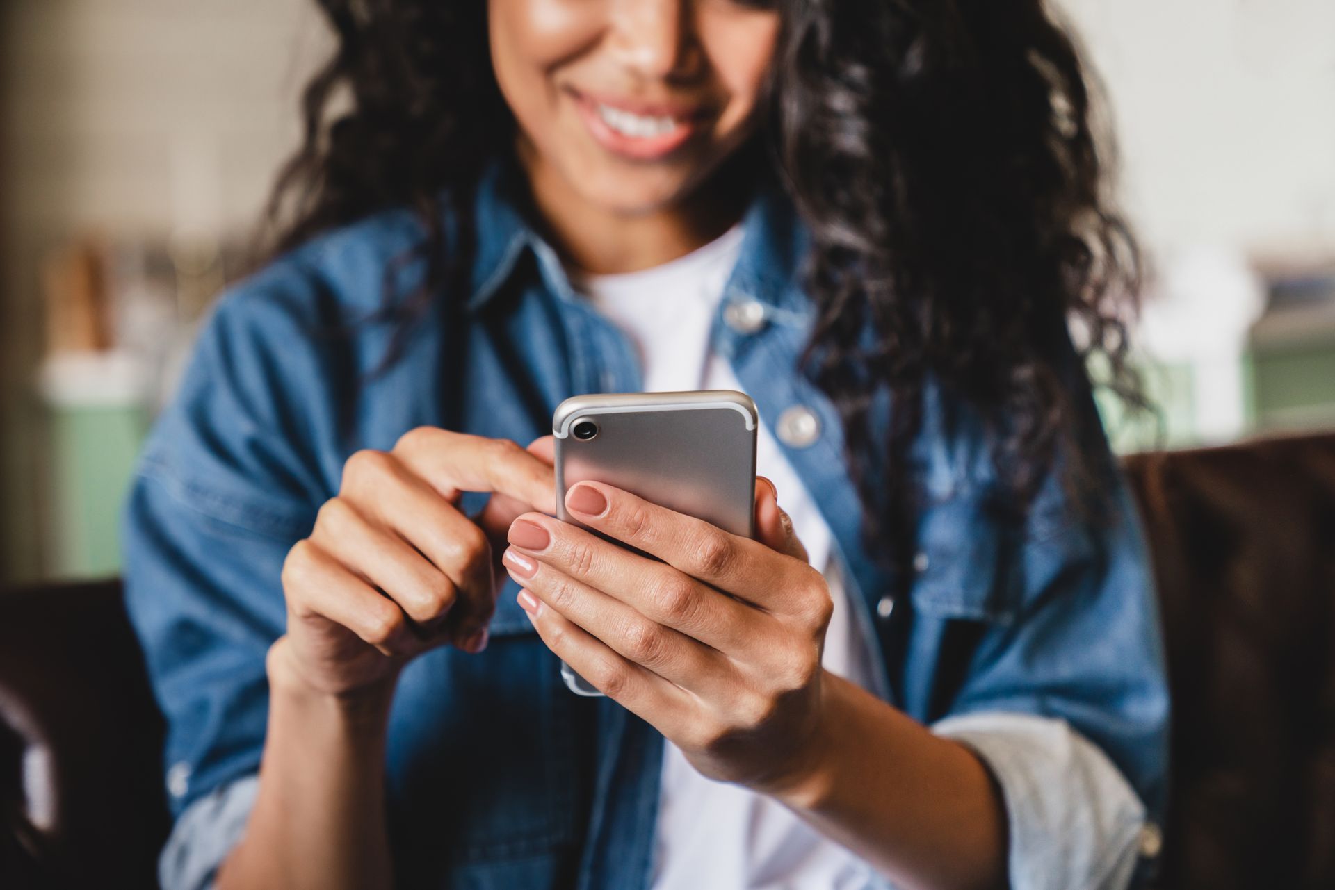 Une personne portant une chemise en jean sourit tout en tenant et en utilisant un smartphone argenté.