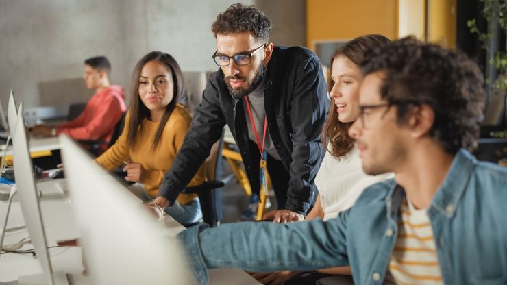 Dans un bureau, un groupe de collègues collabore sur des postes de travail informatiques, l'un d'eux pointant du doigt un écran.