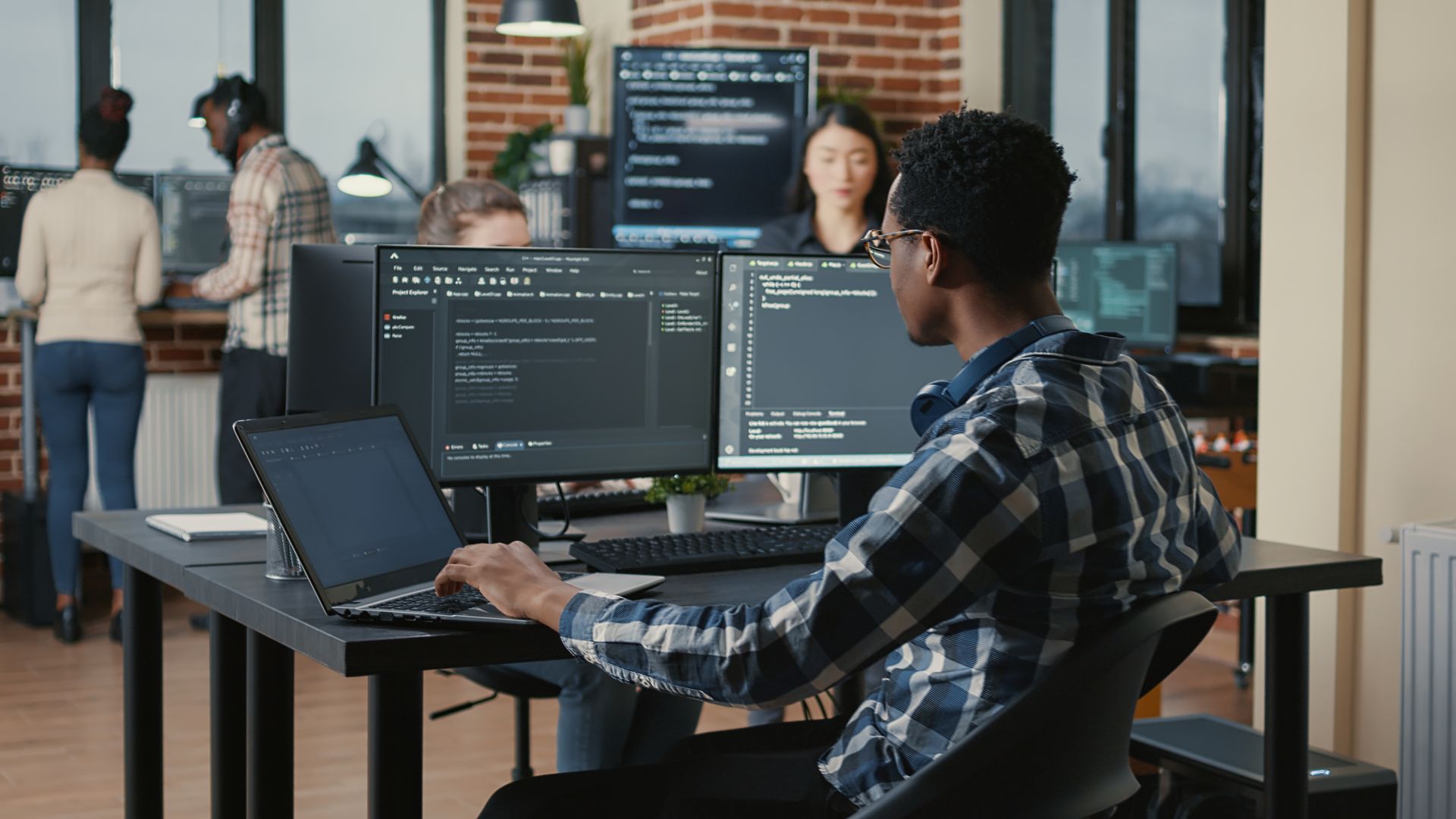 Dans un bureau moderne, un développeur est assis à un bureau équipé de deux écrans et d'un ordinateur portable, en train de taper du code aux côtés de ses collègues.