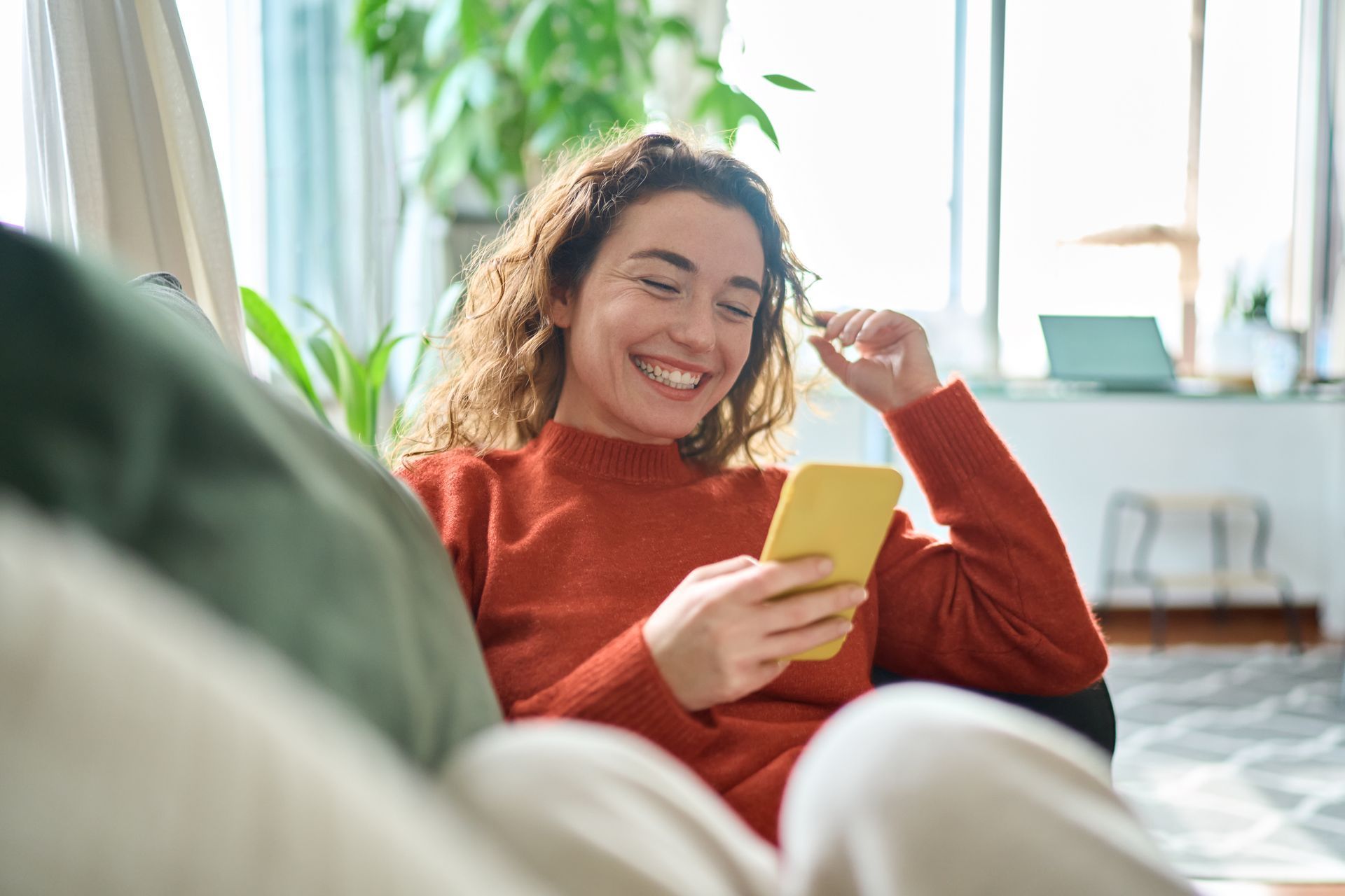 Une personne aux cheveux bouclés sourit, assise sur un canapé, en regardant un smartphone jaune dans un intérieur lumineux.