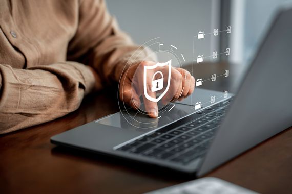 Le doigt d'une personne effleure le pavé tactile d'un ordinateur portable, sur lequel se superposent un bouclier numérique et une icône de cadenas représentant la cybersécurité.
