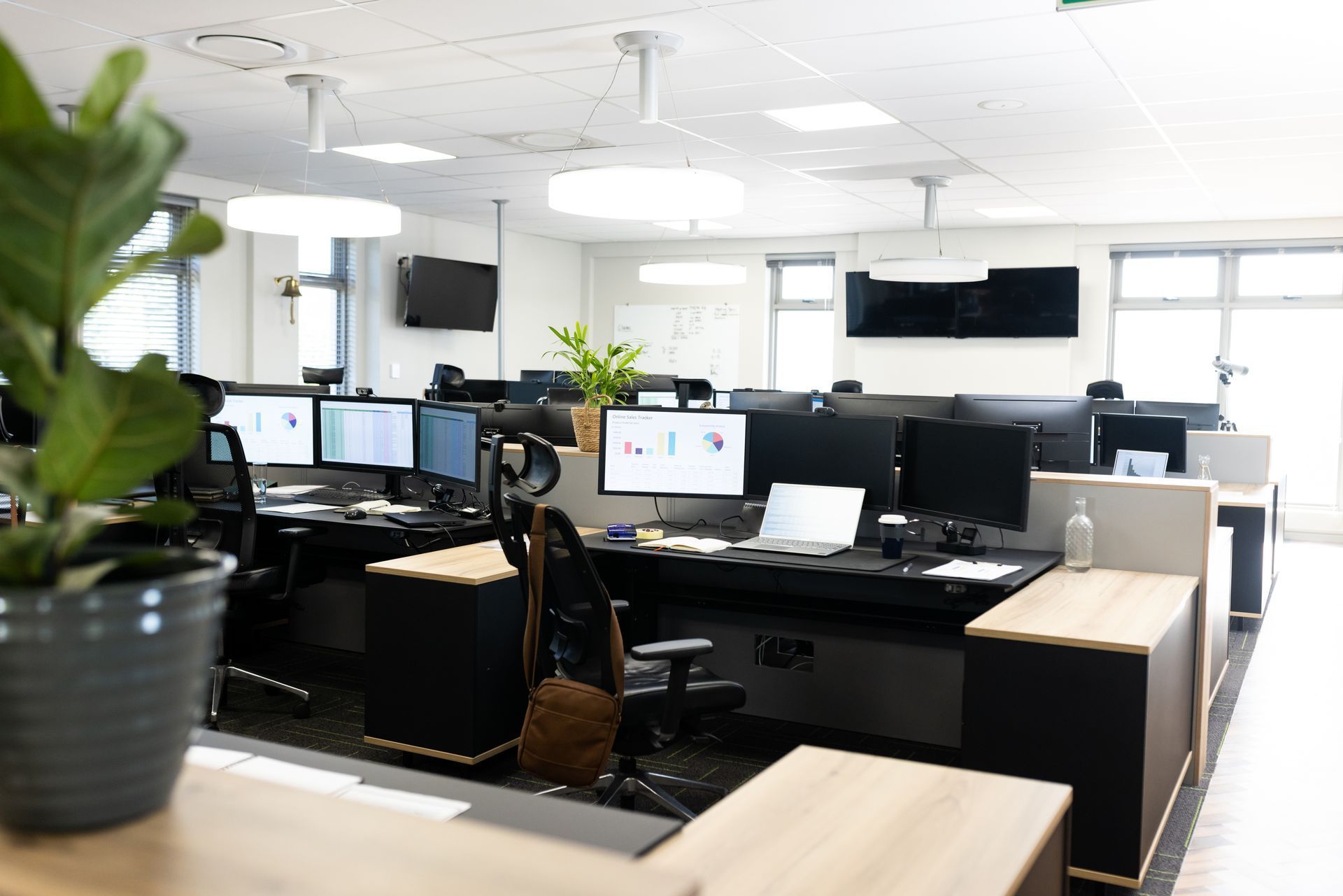 Intérieur de bureau moderne avec des rangées de postes de travail informatiques équipés de plusieurs écrans et une plante en pot au premier plan.