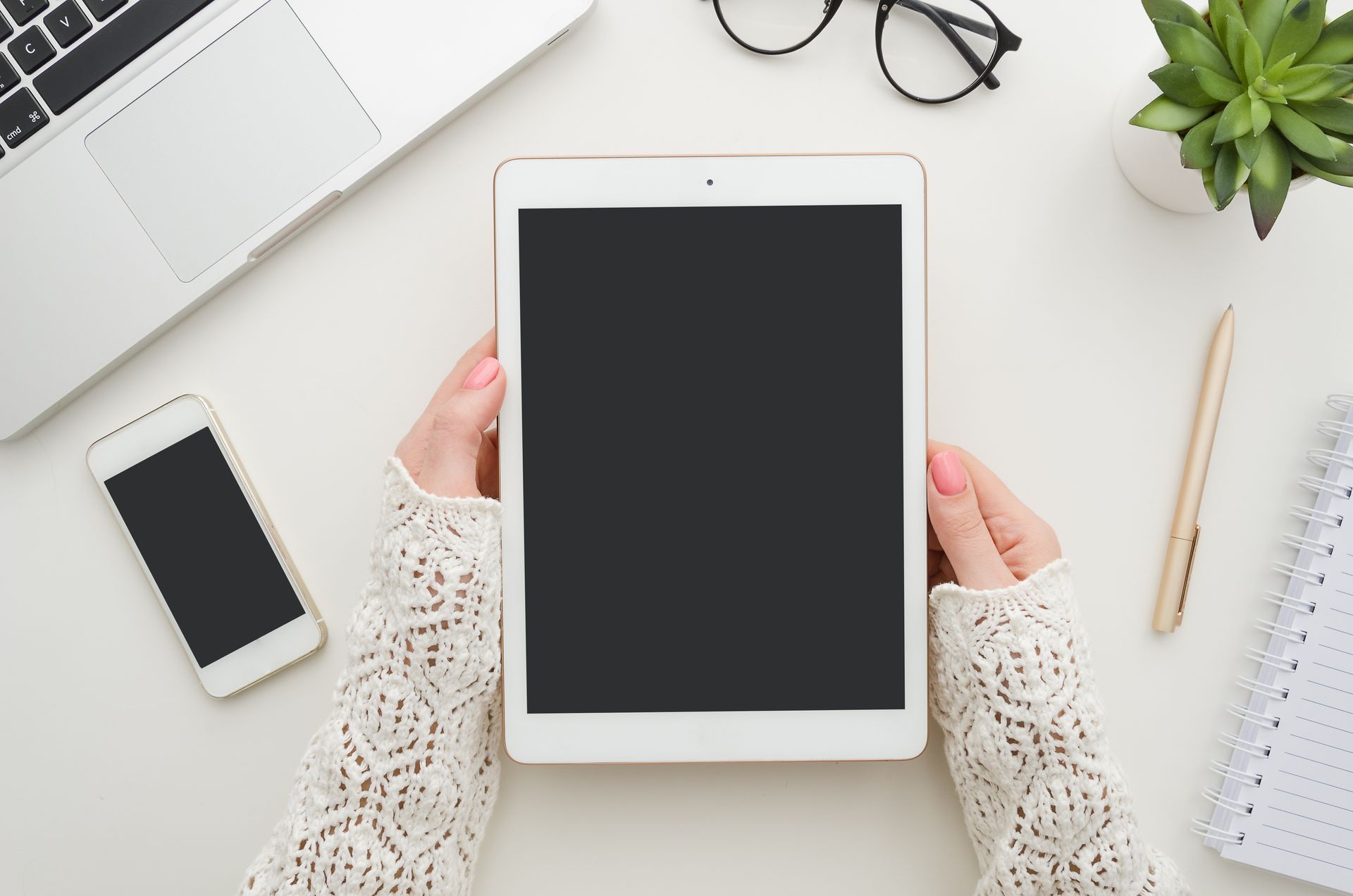 Des mains tiennent une tablette au-dessus d'un bureau blanc où se trouvent également un ordinateur portable, un smartphone, un stylo, des lunettes et une plante grasse.