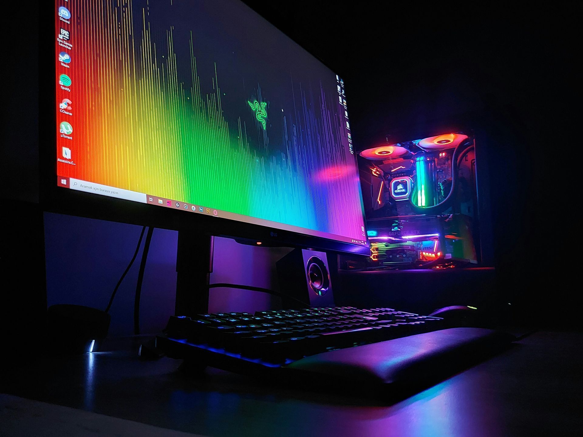 Configuration de bureau gaming rétroéclairée RGB avec écran large, clavier et tour PC lumineuse dans une pièce sombre.