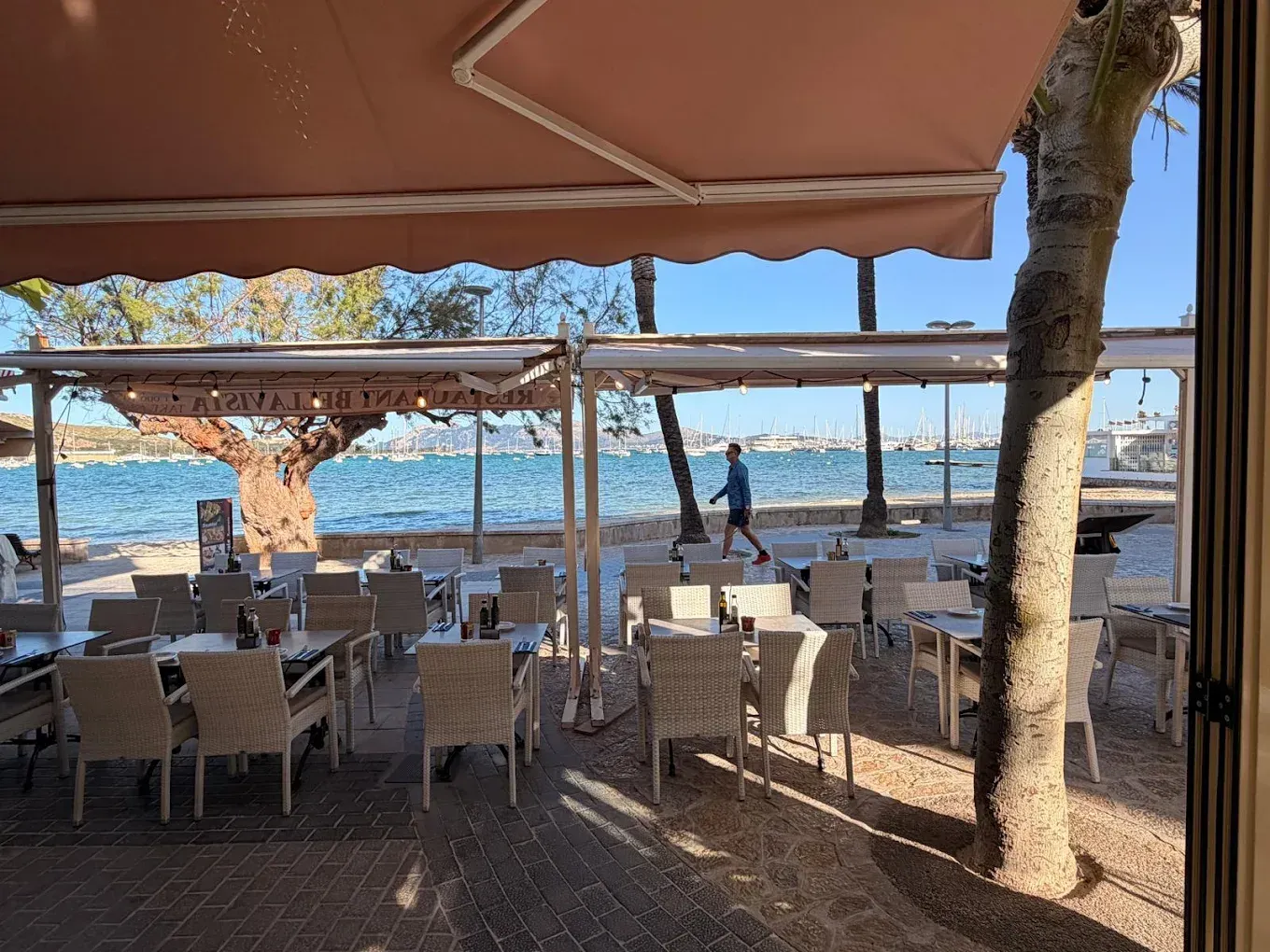 Terraza de un café junto al mar con mesas bajo toldos, con vistas al océano y a la playa al atardecer.