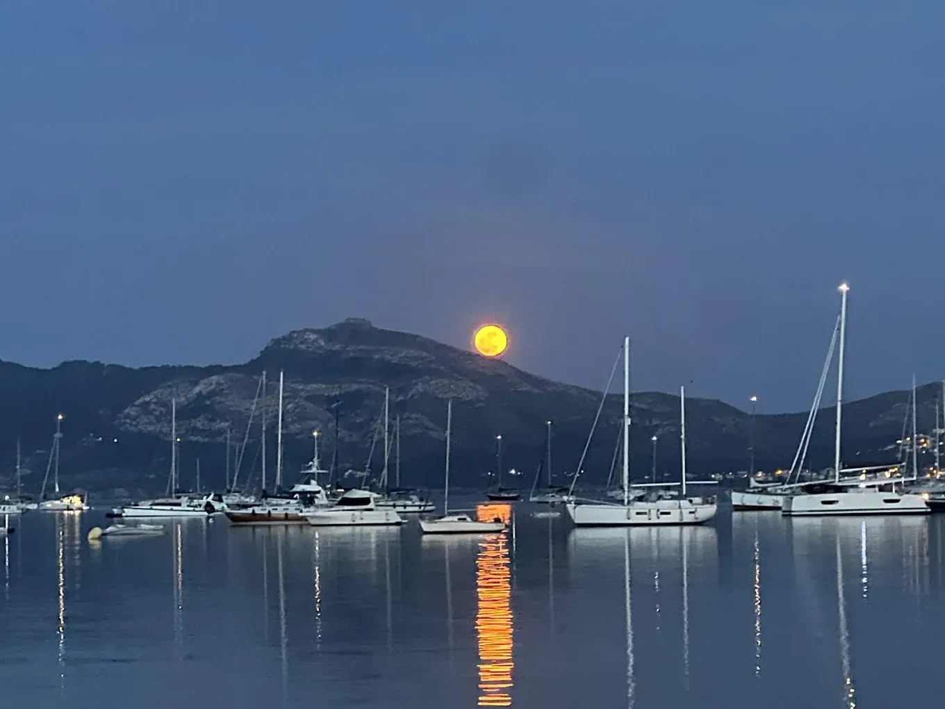 Salida de la luna sobre un puerto deportivo tranquilo con veleros y una ladera oscura al atardecer.