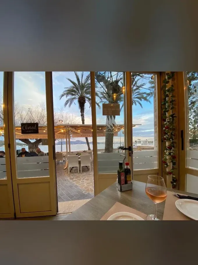 Interior de una cafetería frente a la playa con puertas abiertas, palmeras en el exterior y bebidas en una mesa con vistas al mar.