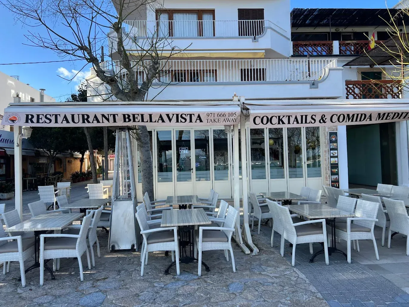 Restaurante al aire libre con sillas y mesas blancas bajo un letrero que dice “Restaurant Bellavista” y “Cocktails & Comida”.