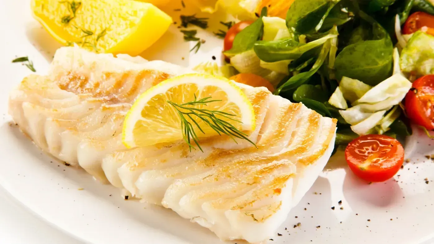 Filete de pescado blanco a la parrilla con limón, eneldo, ensalada verde y tomate cherry en un plato blanco.