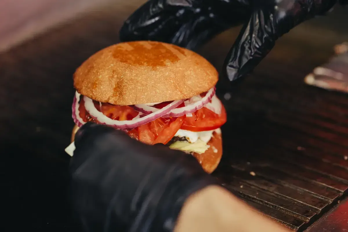 Manos ensamblando una hamburguesa con cebolla roja, tomate y pan, sobre una superficie oscura.