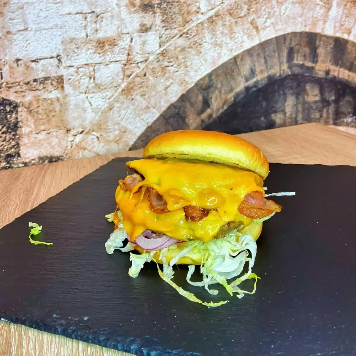 Hamburguesa con queso sobre pizarra negra, con tocino, lechuga y cebolla. Fondo: pared de ladrillos.
