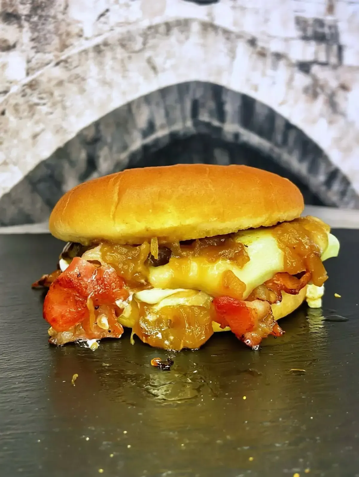 Hamburguesa en pan con queso, tocino y cebolla caramelizada, sobre una superficie oscura, con un fondo de piedra borroso.