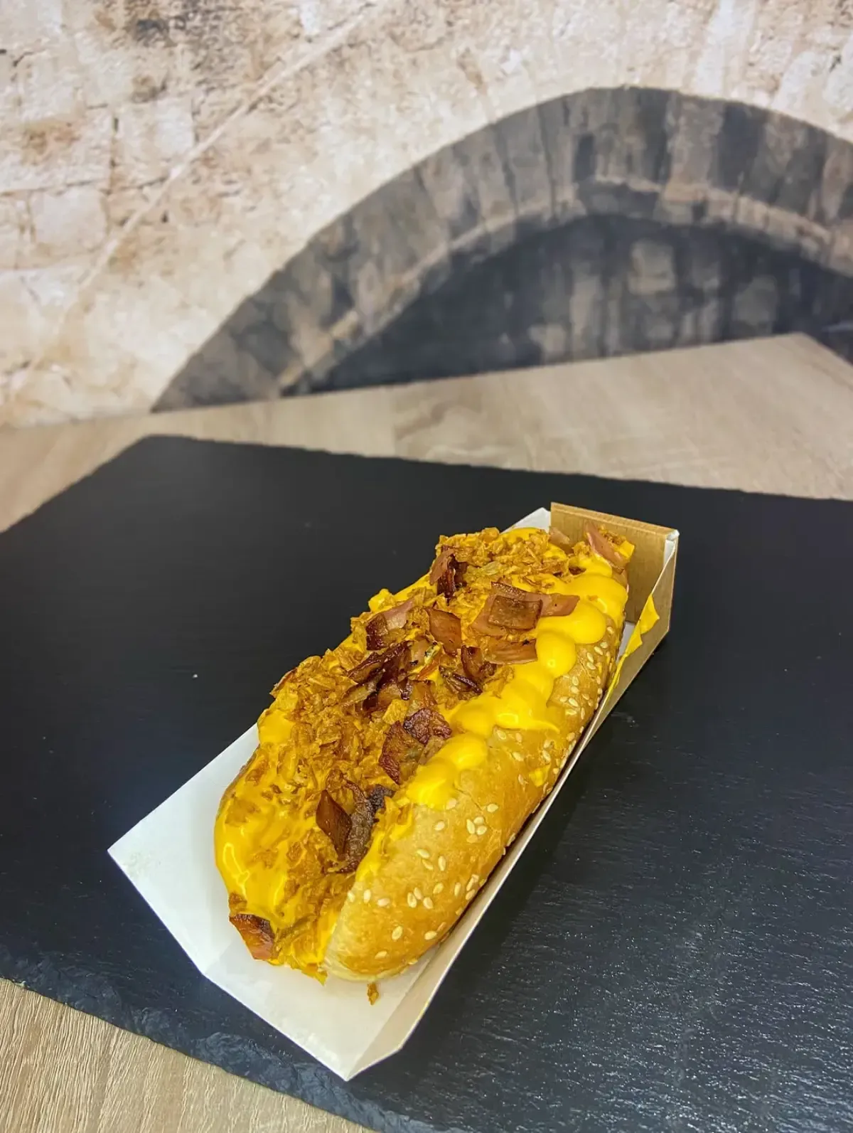 Perro caliente en pan, cubierto con queso y trocitos de tocino, en un recipiente de papel.