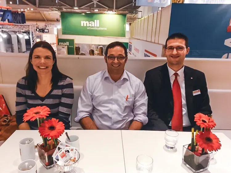 Drei Personen in Business-Kleidung sitzen lächelnd hinter einem Tresen mit roten Blumen auf einer Messe.
