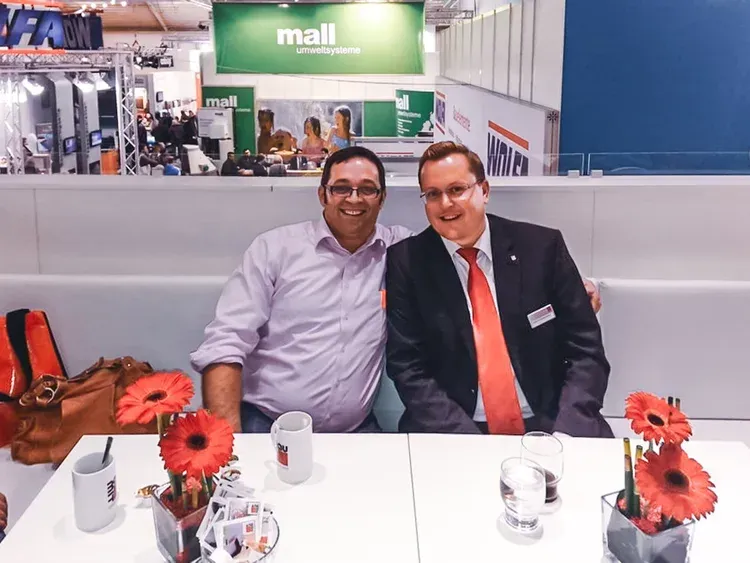 Zwei Männer in geschäftlicher Kleidung sitzen lächelnd an einem Messestand.