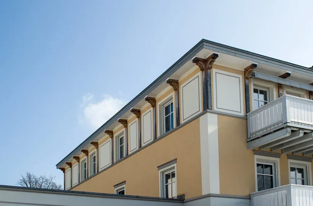 Seitenansicht eines gelb verputzten Gebäudes mit dekorativen weißen Fensterumrandungen und Balkon.