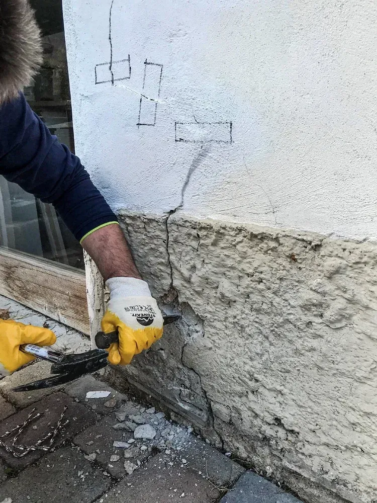 Nahaufnahme einer sanierungsbedürftigen Außenwand mit Rissen und Markierungen am Sockel.