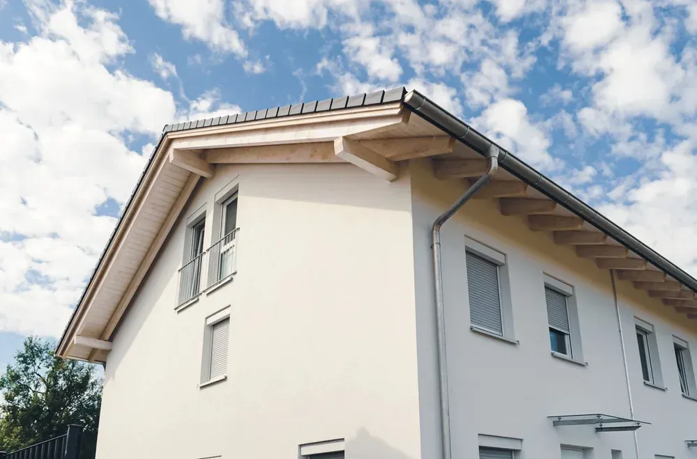 Seitenansicht eines verputzten Hauses mit Dachvorsprung und Regenrinne vor blauem Himmel.