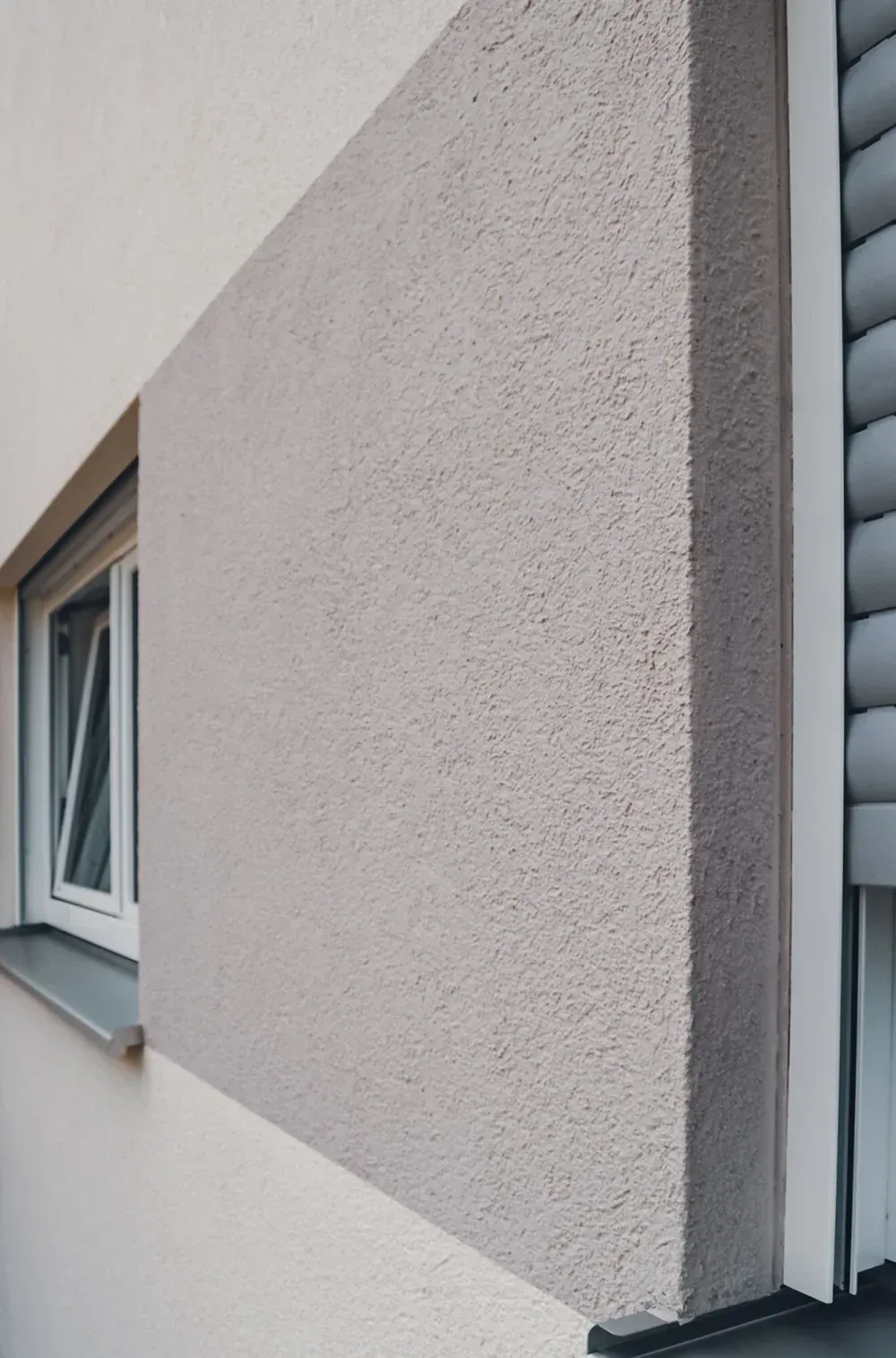 Fassadendetail mit glatter, dunkler Putzfläche, die an eine helle, geriffelte Holz- oder Putzwand grenzt.