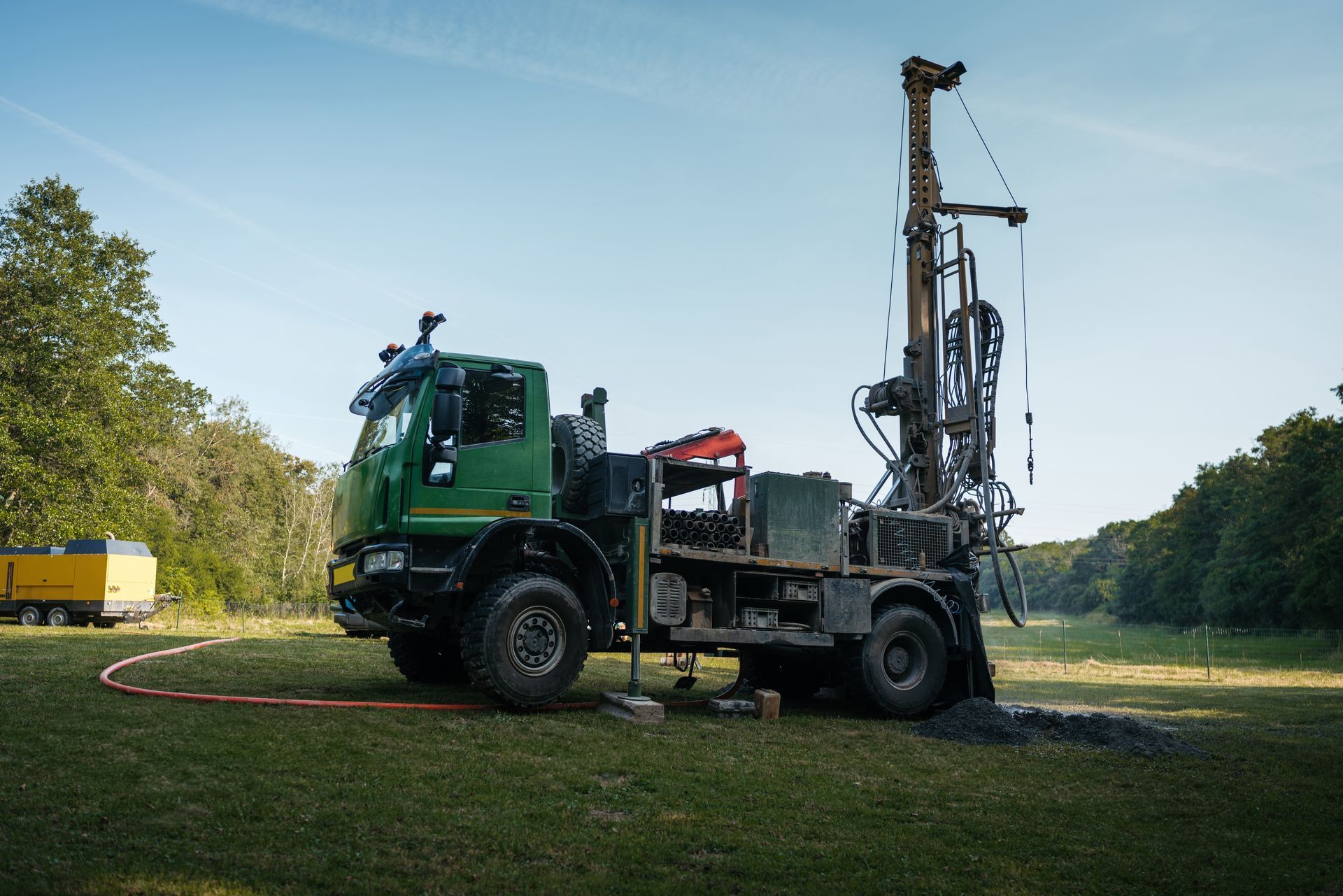 Green drilling rig on a truck in a grassy field, drilling into the ground under a blue sky.