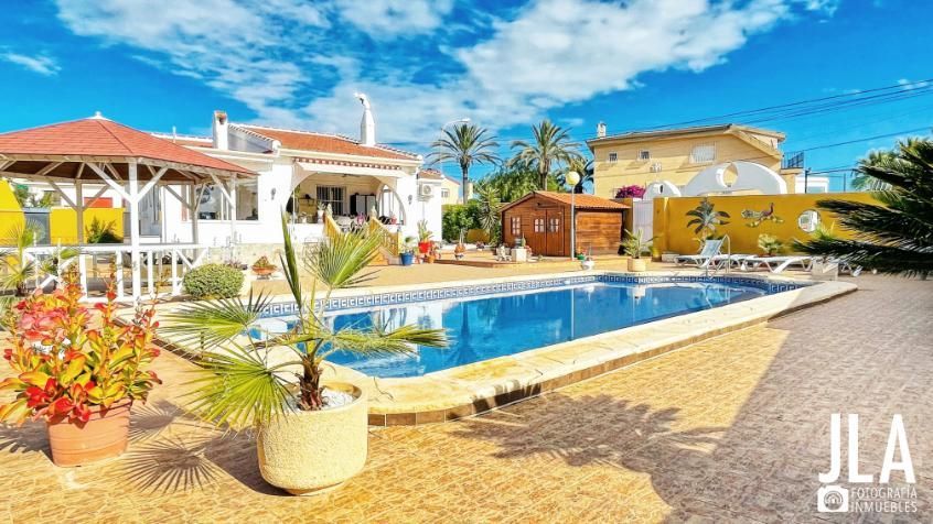 Piscina en un patio trasero soleado con una casa blanca y un cenador.