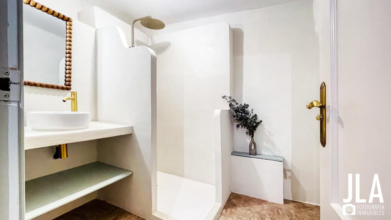 Baño minimalista blanco con accesorios dorados, ducha, lavabo y jarrón decorativo.