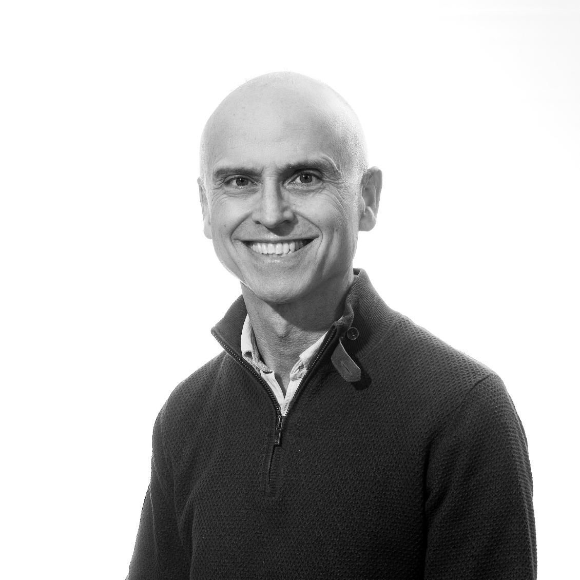 Un hombre calvo que viste un suéter oscuro sonríe, con una camisa de cuello blanco visible sobre un fondo blanco.